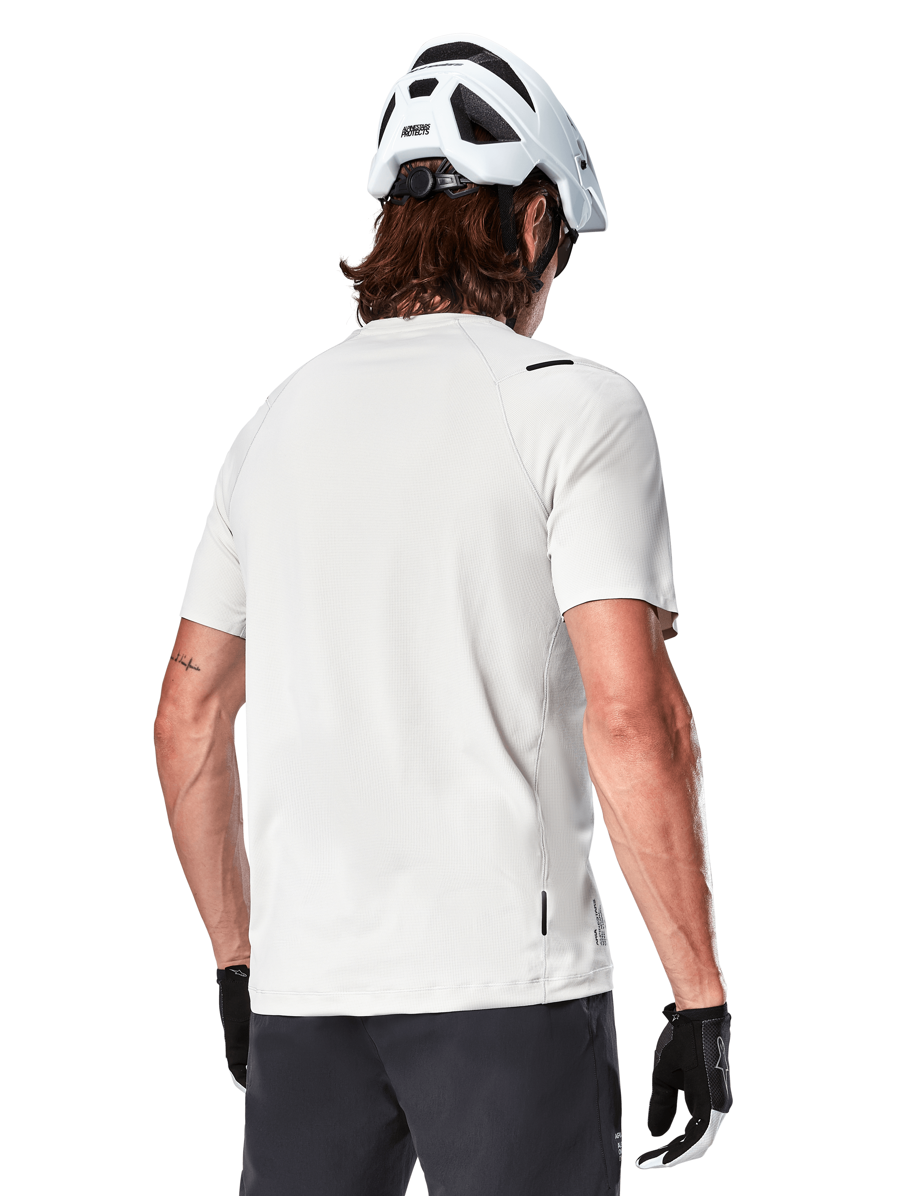 Alpinestars A-Aria Astar Jersey, krótki rękaw, kolor White Dove, widok z tyłu lekkiej i oddychającej koszulki rowerowej MTB, noszonej z białym kaskiem i czarnymi rękawiczkami rowerowymi, z wydłużonym tyłem dla lepszej osłony