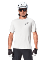 Rowerzysta w koszulce Alpinestars A-Aria Astar z krótkim rękawem, kolor White Dove z małym czarnym logo Astar na piersi, biały kask rowerowy, czarne okulary przeciwsłoneczne i czarno-białe rękawiczki, profesjonalna odzież MTB do jazdy w terenie
