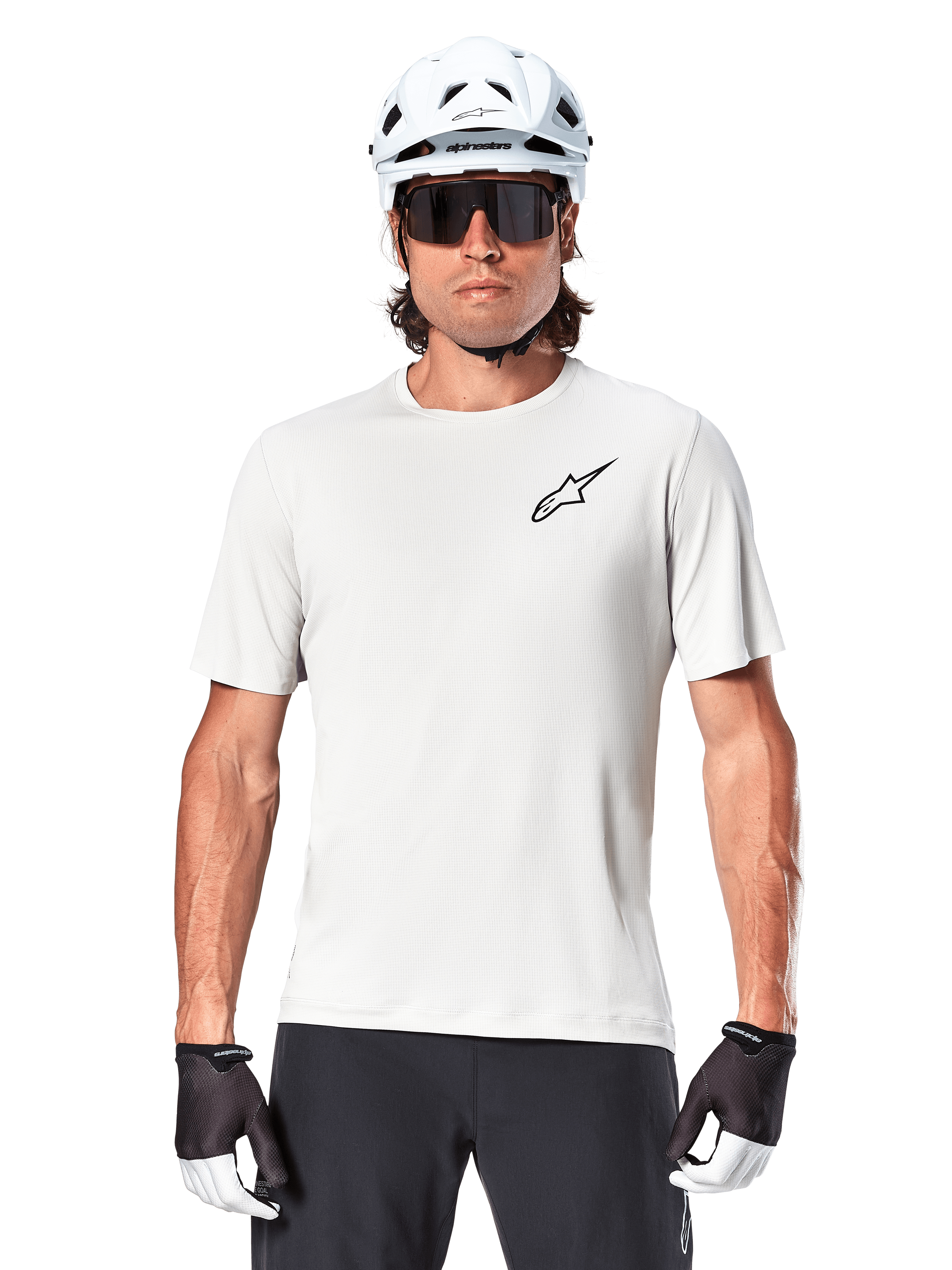 Rowerzysta w koszulce Alpinestars A-Aria Astar z krótkim rękawem, kolor White Dove z małym czarnym logo Astar na piersi, biały kask rowerowy, czarne okulary przeciwsłoneczne i czarno-białe rękawiczki, profesjonalna odzież MTB do jazdy w terenie