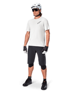 Rowerzysta MTB w koszulce Alpinestars A-Aria Astar z krótkim rękawem w kolorze White Dove, czarne szorty rowerowe, biały kask i czarne ochraniacze na kolana, małe czarne logo na piersi, dopasowany zestaw odzieży rowerowej do jazdy po szlakach