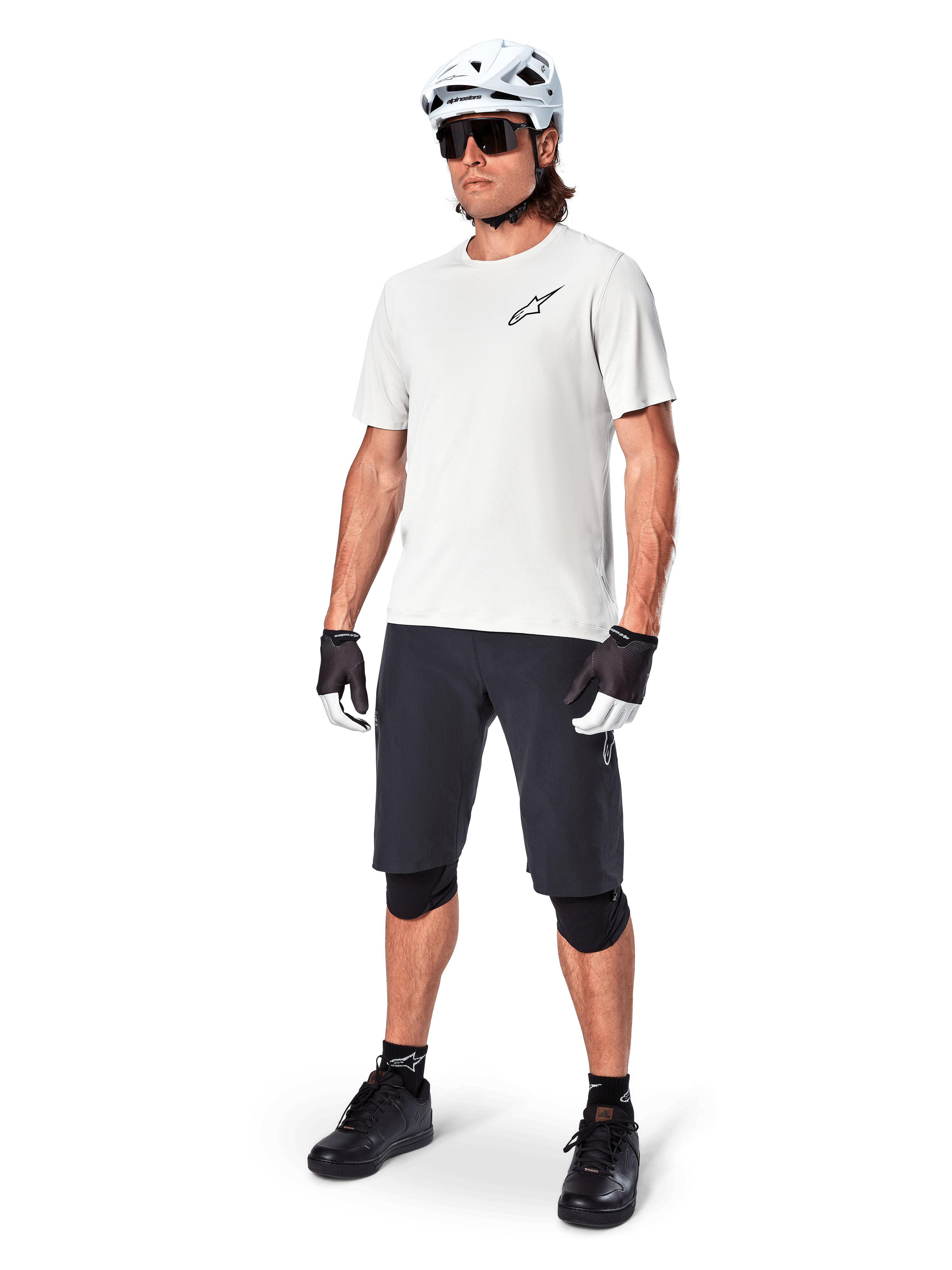 Rowerzysta MTB w koszulce Alpinestars A-Aria Astar z krótkim rękawem w kolorze White Dove, czarne szorty rowerowe, biały kask i czarne ochraniacze na kolana, małe czarne logo na piersi, dopasowany zestaw odzieży rowerowej do jazdy po szlakach