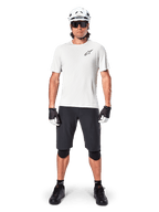 Rowerzysta ubrany w koszulkę Alpinestars A-Aria Astar z krótkim rękawem w kolorze White Dove, czarne logo Astars na piersi, czarne szorty MTB, biały kask, okulary przeciwsłoneczne, czarne ochraniacze kolan i rękawiczki, kompletny zestaw do jazdy w terenie.