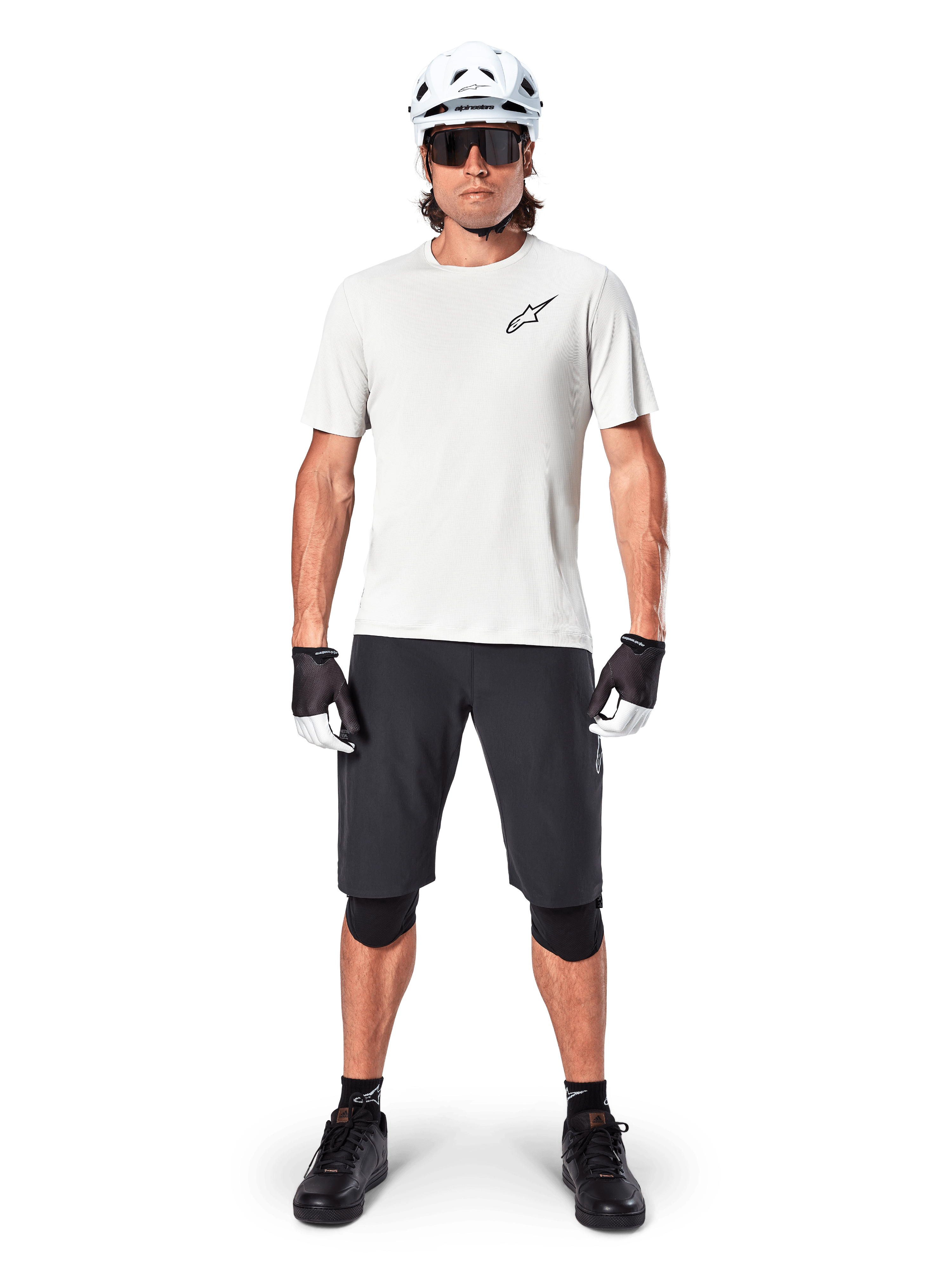 Rowerzysta ubrany w koszulkę Alpinestars A-Aria Astar z krótkim rękawem w kolorze White Dove, czarne logo Astars na piersi, czarne szorty MTB, biały kask, okulary przeciwsłoneczne, czarne ochraniacze kolan i rękawiczki, kompletny zestaw do jazdy w terenie.
