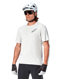 Alpinestars A-Aria Astar Short Sleeve Jersey, kolor White Dove, koszulka noszona przez rowerzystę w białym kasku i ciemnych okularach przeciwsłonecznych, małe czarne logo gwiazdy na piersi, oddychająca odzież wyczynowa do jazdy MTB