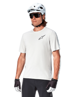 Alpinestars A-Aria Astar Short Sleeve Jersey, kolor White Dove, koszulka noszona przez rowerzystę w białym kasku i ciemnych okularach przeciwsłonecznych, małe czarne logo gwiazdy na piersi, oddychająca odzież wyczynowa do jazdy MTB