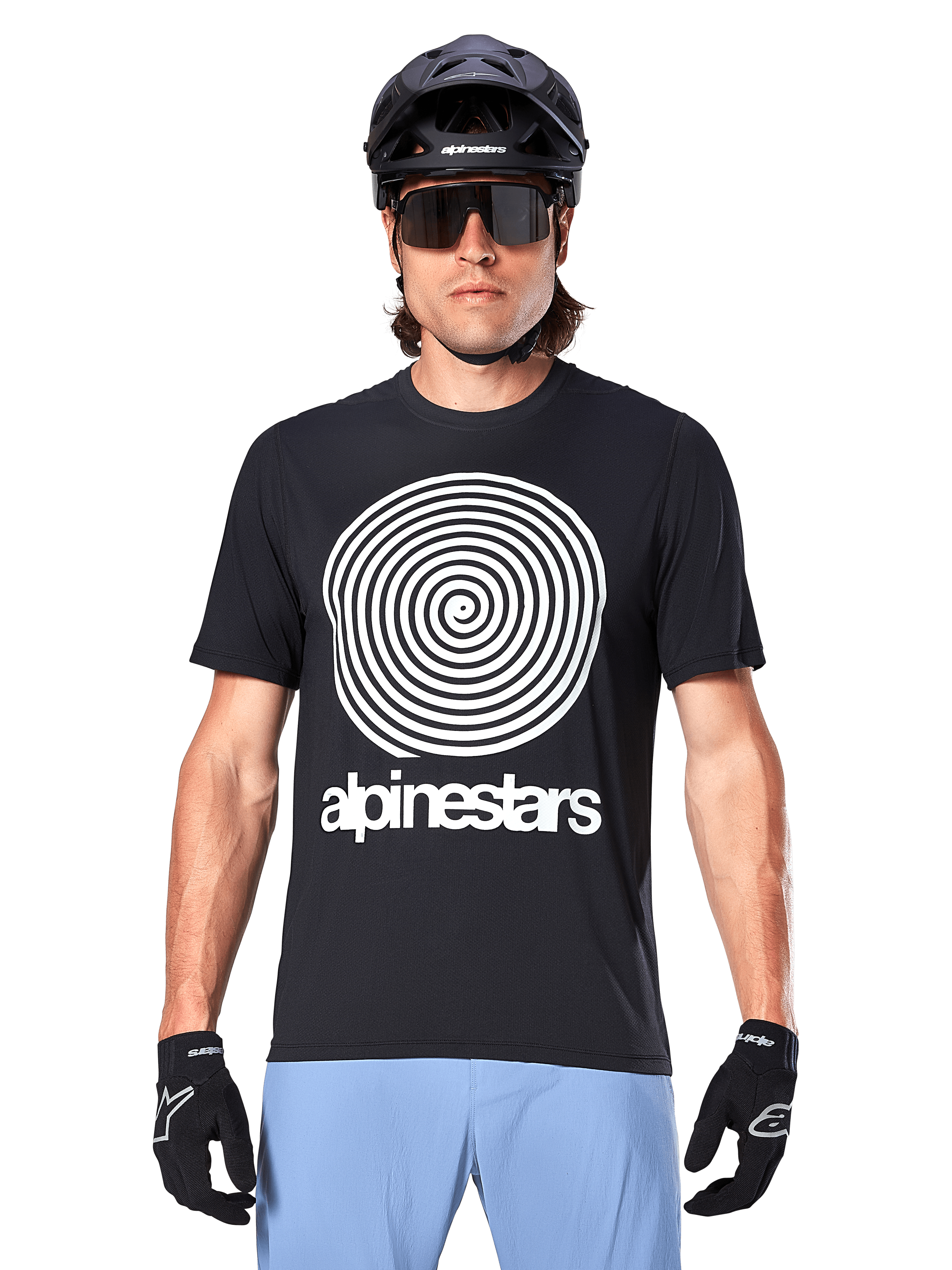 Koszulka Alpinestars A-Dura Oscar, koszulka MTB z krótkim rękawem, czarna, z dużą białą spiralną grafiką Oscar w stylu vintage na piersi, noszona z czarnym kaskiem rowerowym, okularami przeciwsłonecznymi, czarnymi rękawiczkami i jasnoniebieskimi szortami, odzież rowerowa off-road inspirowana dziedzictwem