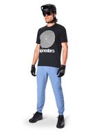 Alpinestars A-Dura Oscar Short Sleeve Jersey, czarna koszulka MTB z dużą białą spiralną grafiką heritage, noszona przez kolarza w czarnym kasku, okularach przeciwsłonecznych, czarnych rękawiczkach i jasnoniebieskich spodniach, profesjonalna odzież do kolarstwa górskiego