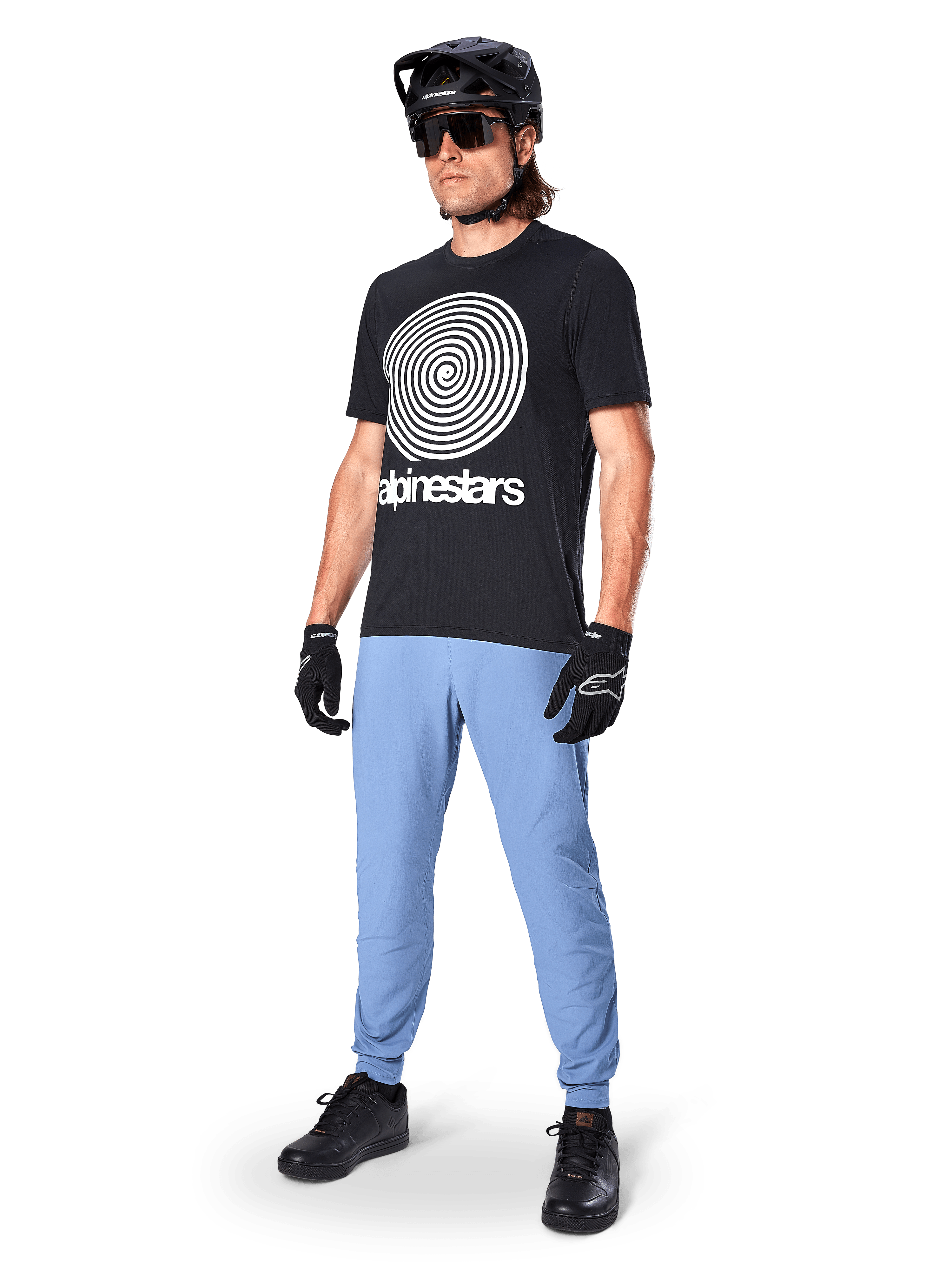 Alpinestars A-Dura Oscar Short Sleeve Jersey, czarna koszulka MTB z dużą białą spiralną grafiką heritage, noszona przez kolarza w czarnym kasku, okularach przeciwsłonecznych, czarnych rękawiczkach i jasnoniebieskich spodniach, profesjonalna odzież do kolarstwa górskiego
