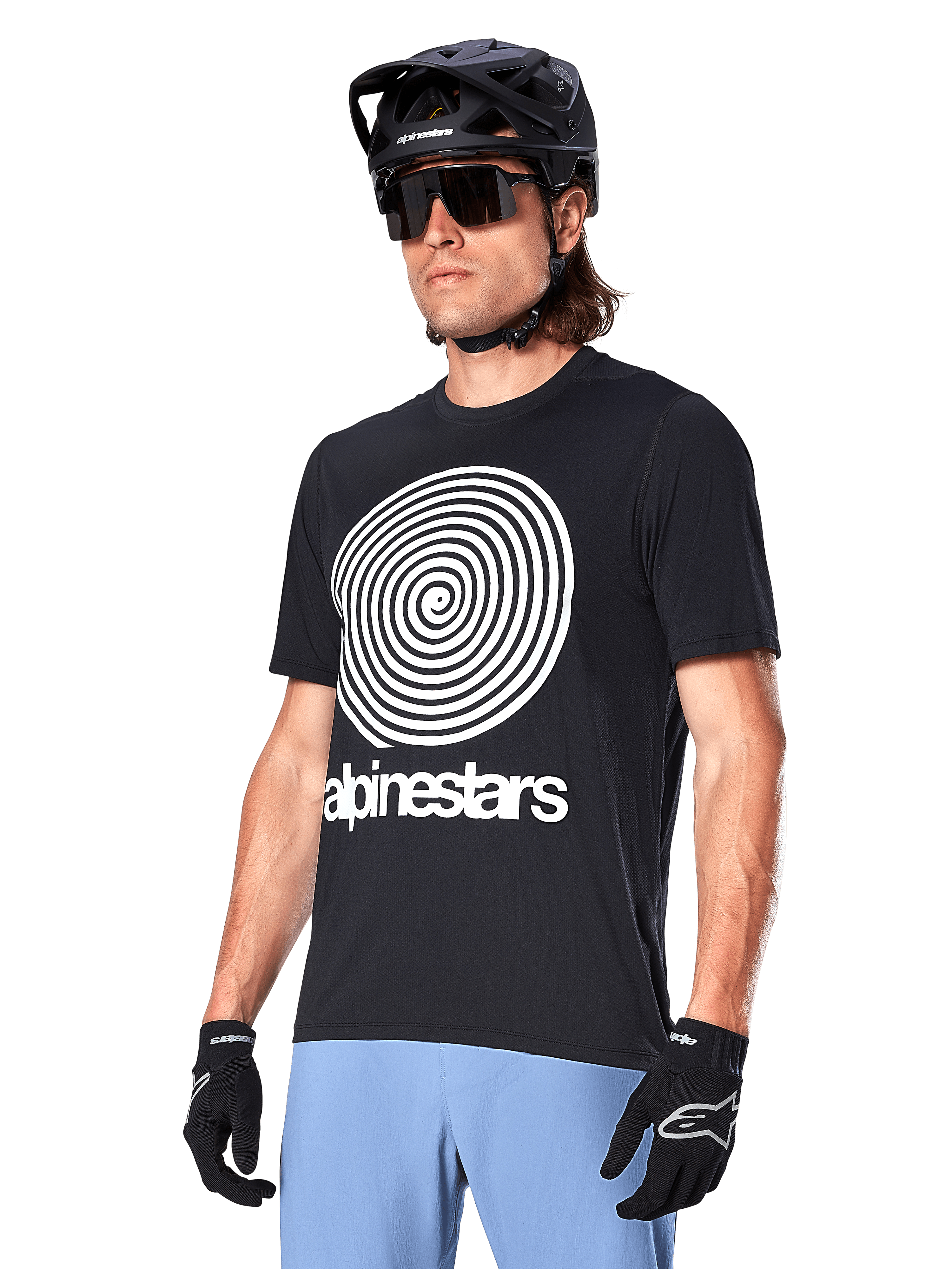 Koszulka Alpinestars A-Dura Oscar - krótki rękaw, czarna, z dużą białą spiralną grafiką i logo Alpinestars na piersi, kolarz w czarnym kasku MTB, okularach przeciwsłonecznych i jasnoniebieskich spodniach