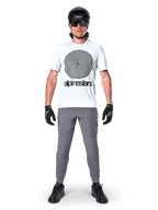 Rowerzysta górski w koszulce Alpinestars A-Dura Oscar z krótkim rękawem, kolor White Dove z dużym czarnym spiralnym logo, szare spodnie, czarny kask, okulary przeciwsłoneczne i czarne rękawiczki, wyczynowy strój MTB do jazdy trailowej