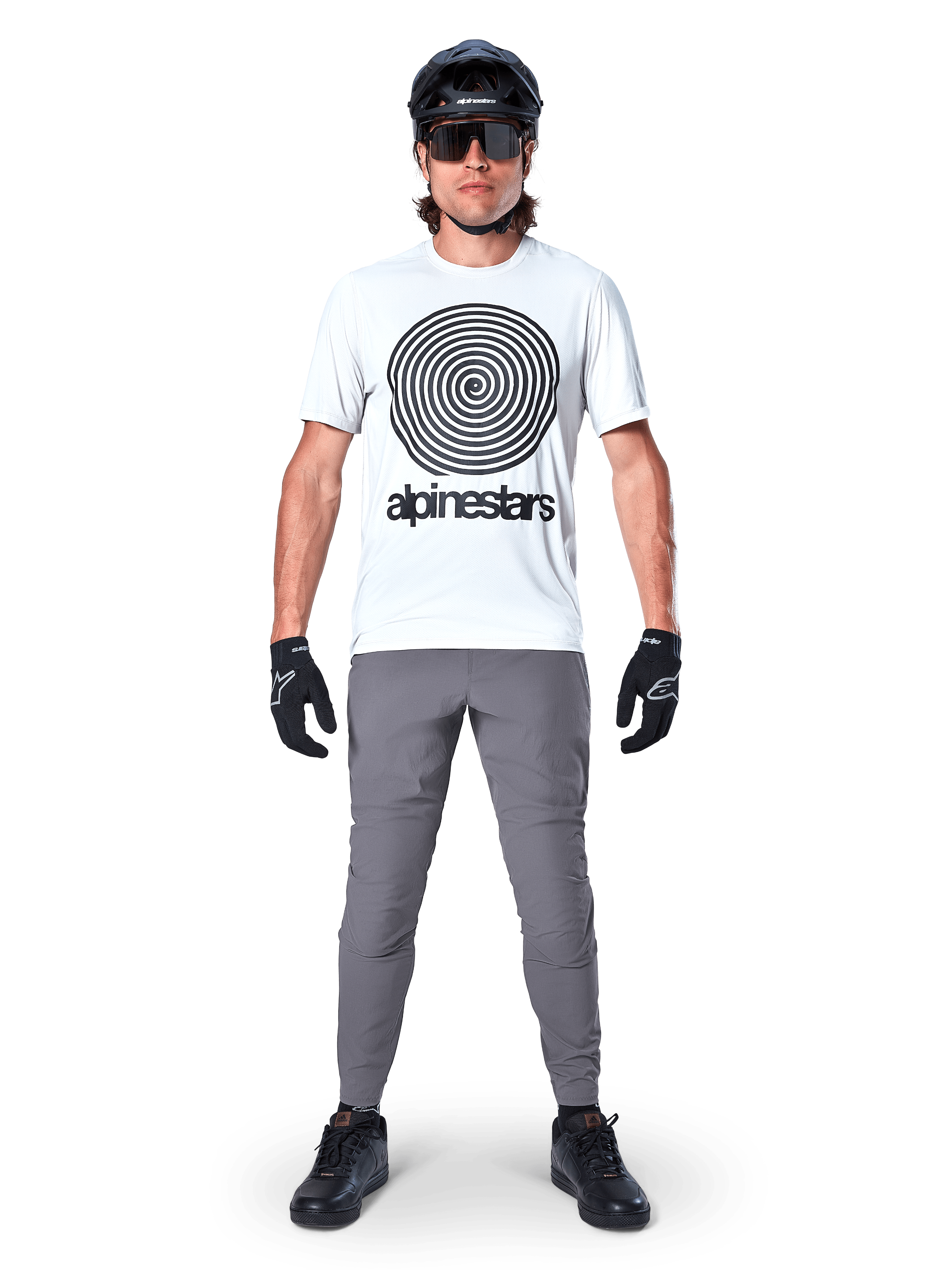 Rowerzysta górski w koszulce Alpinestars A-Dura Oscar z krótkim rękawem, kolor White Dove z dużym czarnym spiralnym logo, szare spodnie, czarny kask, okulary przeciwsłoneczne i czarne rękawiczki, wyczynowy strój MTB do jazdy trailowej