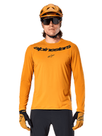 Koszulka z długim rękawem Alpinestars A-Dura Rocker w kolorze ciemnozłotym, z czarnym logotypem na piersi, noszona z pasującym złotym kaskiem i rękawicami do jazdy na rowerze górskim.