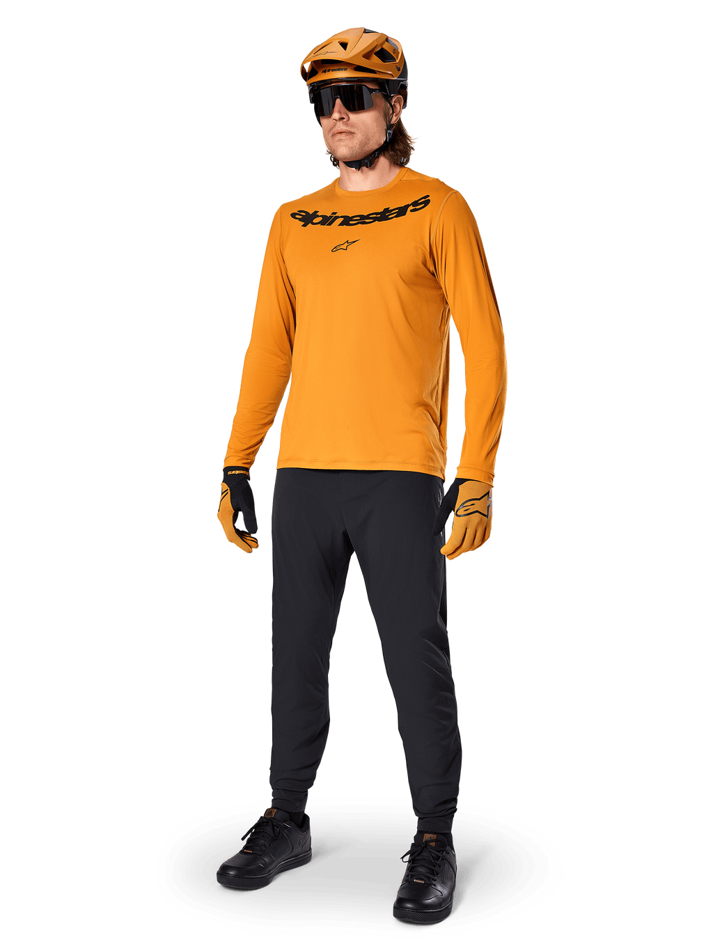 Koszulka z długim rękawem Alpinestars A-Dura Rocker, kolarska koszulka w kolorze Dark Gold, noszona z dopasowanym kaskiem MTB i czarnymi spodniami, z czarnym logotypem na klatce piersiowej, skoordynowana odzież wyczynowa do jazdy terenowej na rowerze górskim