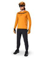 Koszulka z długim rękawem Alpinestars A-Dura Rocker, kolarska koszulka w kolorze Dark Gold, noszona z dopasowanym kaskiem MTB i czarnymi spodniami, z czarnym logotypem na klatce piersiowej, skoordynowana odzież wyczynowa do jazdy terenowej na rowerze górskim