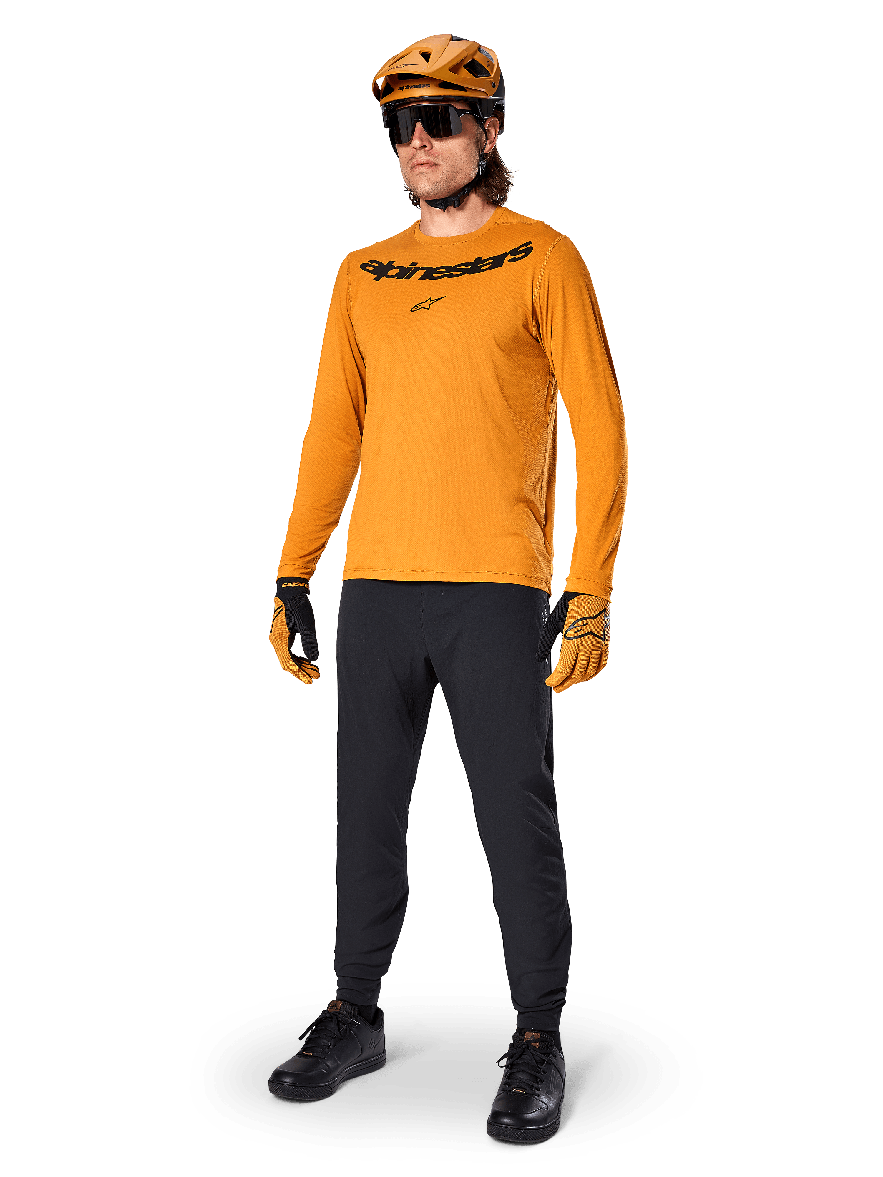 Koszulka z długim rękawem Alpinestars A-Dura Rocker, kolarska koszulka w kolorze Dark Gold, noszona z dopasowanym kaskiem MTB i czarnymi spodniami, z czarnym logotypem na klatce piersiowej, skoordynowana odzież wyczynowa do jazdy terenowej na rowerze górskim