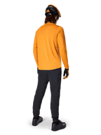 Rowerzysta górski widziany od tyłu, ubrany w koszulkę Alpinestars A-Dura Rocker Long Sleeve w kolorze Dark Gold, czarne spodnie, pomarańczowo-czarny kask i czarne buty, profesjonalny zestaw odzieży MTB do jazdy po szlakach
