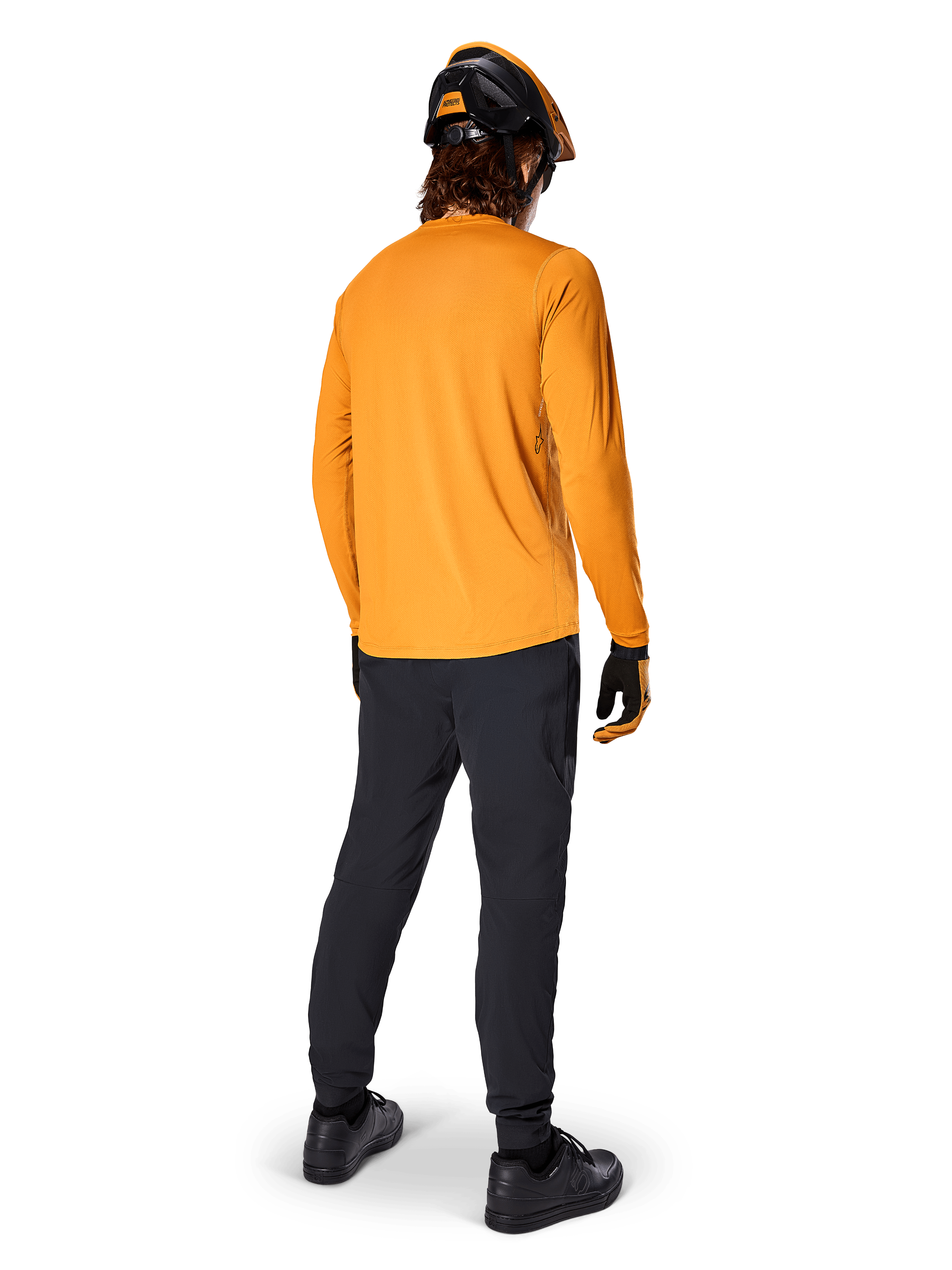 Rowerzysta górski widziany od tyłu, ubrany w koszulkę Alpinestars A-Dura Rocker Long Sleeve w kolorze Dark Gold, czarne spodnie, pomarańczowo-czarny kask i czarne buty, profesjonalny zestaw odzieży MTB do jazdy po szlakach
