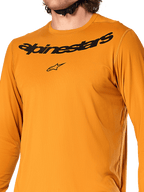 Alpinestars A-Dura Rocker Long Sleeve Jersey, kolor ciemnozłoty, zbliżenie na klatkę piersiową z czarnym napisem Alpinestars i logo gwiazdy, teksturowany dżakardowy materiał odprowadzający wilgoć, techniczna odzież rowerowa MTB