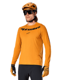 Alpinestars A-Dura Rocker Long Sleeve Jersey, koszulka rowerowa MTB, kolor ciemnozłoty, osoba w koszulce z dopasowanym kaskiem i rękawiczkami, sportowy spójny zestaw z napisem Alpinestars na piersi