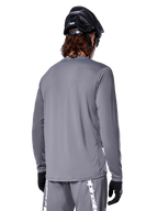 Alpinestars A-Dura Rocker Long Sleeve Jersey, ciemnoszara koszulka rowerowa z długim rękawem, widok kolarza górskiego od tyłu, pasujące szare spodenki z białym logo, czarny kask MTB i czarne rękawiczki, profesjonalna odzież do kolarstwa terenowego