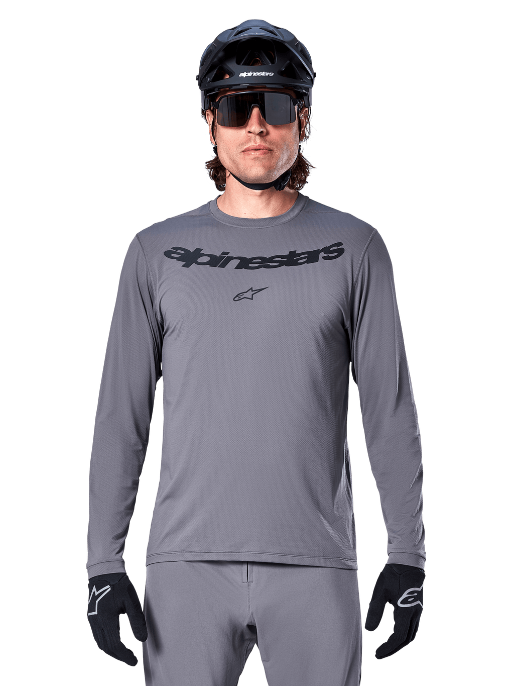 Mężczyzna kolarz w koszulce Alpinestars A-Dura Rocker z długim rękawem, ciemnoszara koszulka MTB z czarnym napisem na piersi, szare spodnie, czarny kask i rękawice, strój do jazdy terenowej