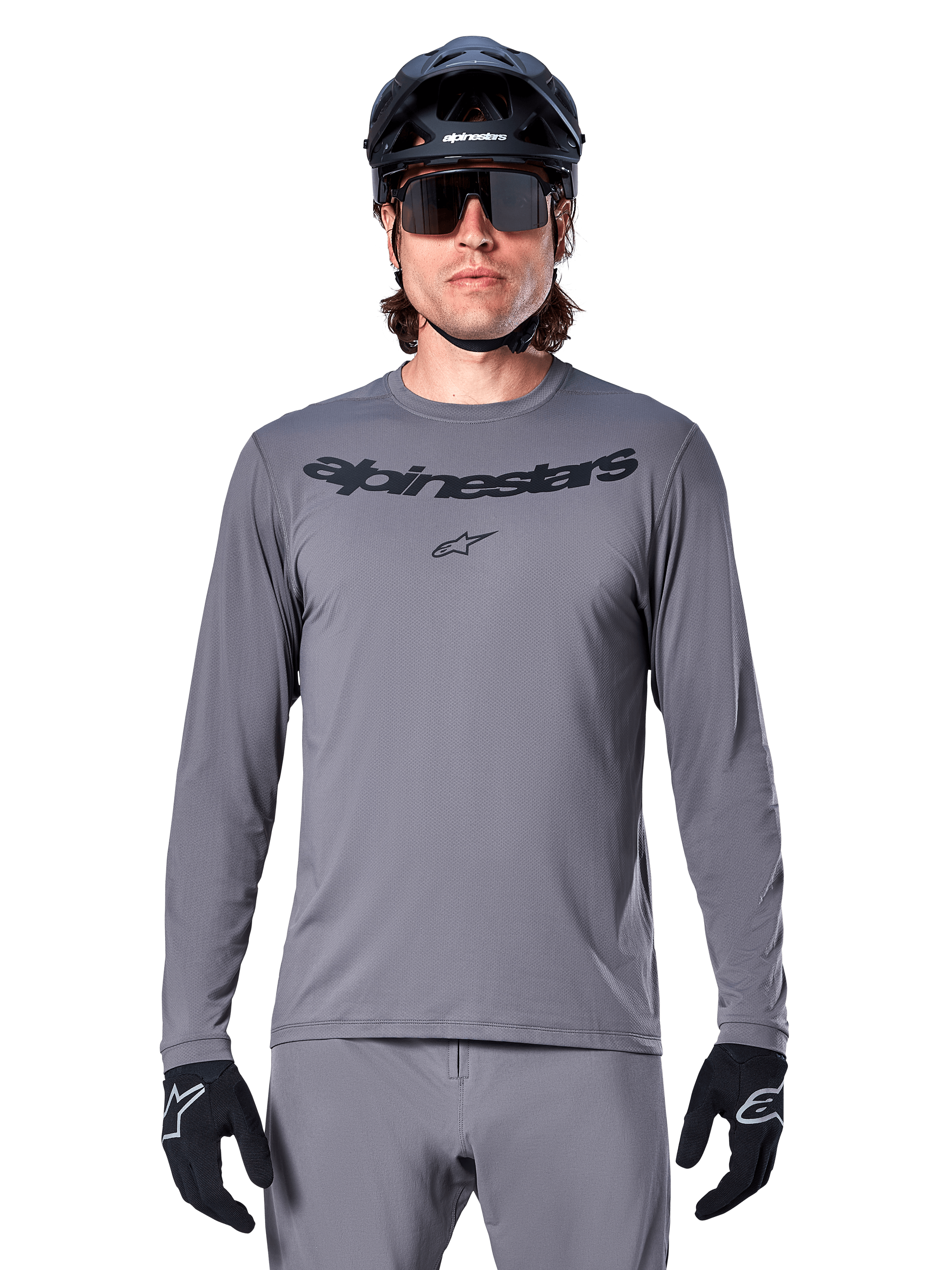 Mężczyzna kolarz w koszulce Alpinestars A-Dura Rocker z długim rękawem, ciemnoszara koszulka MTB z czarnym napisem na piersi, szare spodnie, czarny kask i rękawice, strój do jazdy terenowej