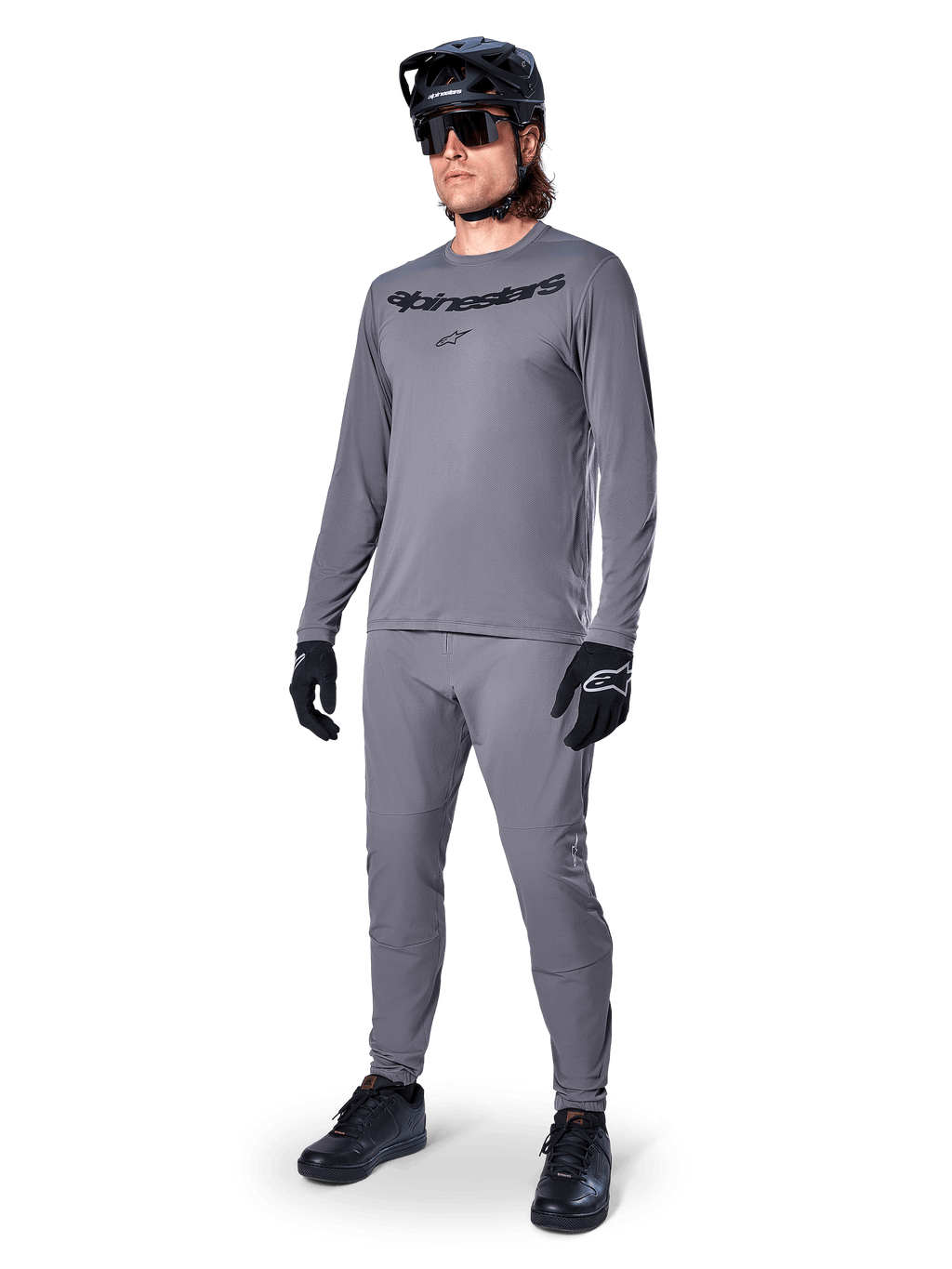 Koszulka Alpinestars A-Dura Rocker z długim rękawem, ciemnoszara, noszona przez modela z dopasowanymi szarymi spodniami, czarnym kaskiem MTB, okularami przeciwsłonecznymi i rękawicami, swobodny styl MTB do jazdy w terenie