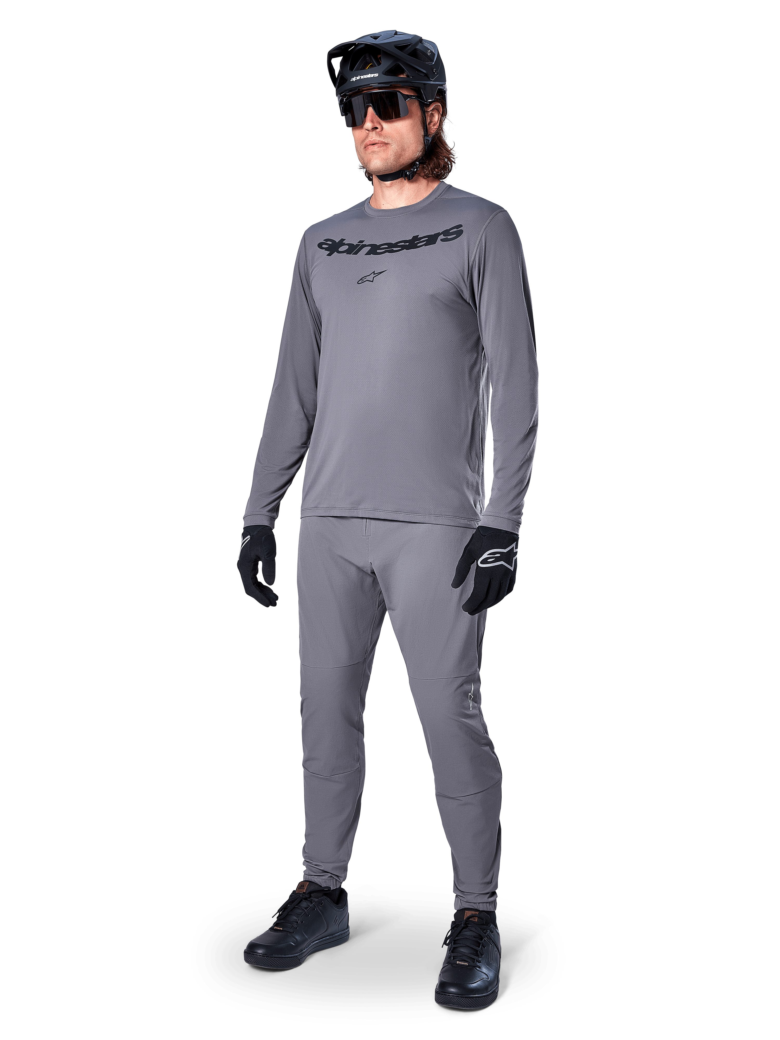 Koszulka Alpinestars A-Dura Rocker z długim rękawem, ciemnoszara, noszona przez modela z dopasowanymi szarymi spodniami, czarnym kaskiem MTB, okularami przeciwsłonecznymi i rękawicami, swobodny styl MTB do jazdy w terenie