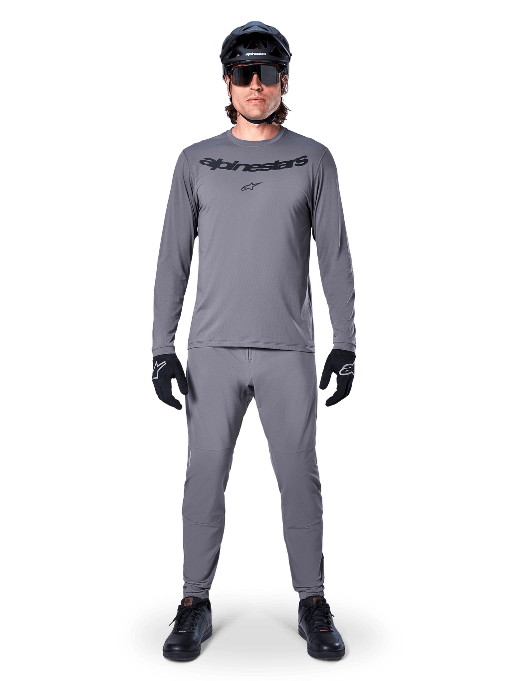 Kolarz w koszulce Alpinestars A-Dura Rocker z długim rękawem w kolorze ciemnoszarym, pasujące szare spodnie, czarny kask MTB, okulary przeciwsłoneczne i rękawiczki, profesjonalna odzież do jazdy na rowerze górskim na szlaki terenowe