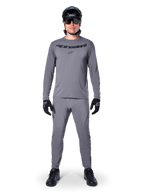 Kolarz w koszulce Alpinestars A-Dura Rocker z długim rękawem w kolorze ciemnoszarym, pasujące szare spodnie, czarny kask MTB, okulary przeciwsłoneczne i rękawiczki, profesjonalna odzież do jazdy na rowerze górskim na szlaki terenowe