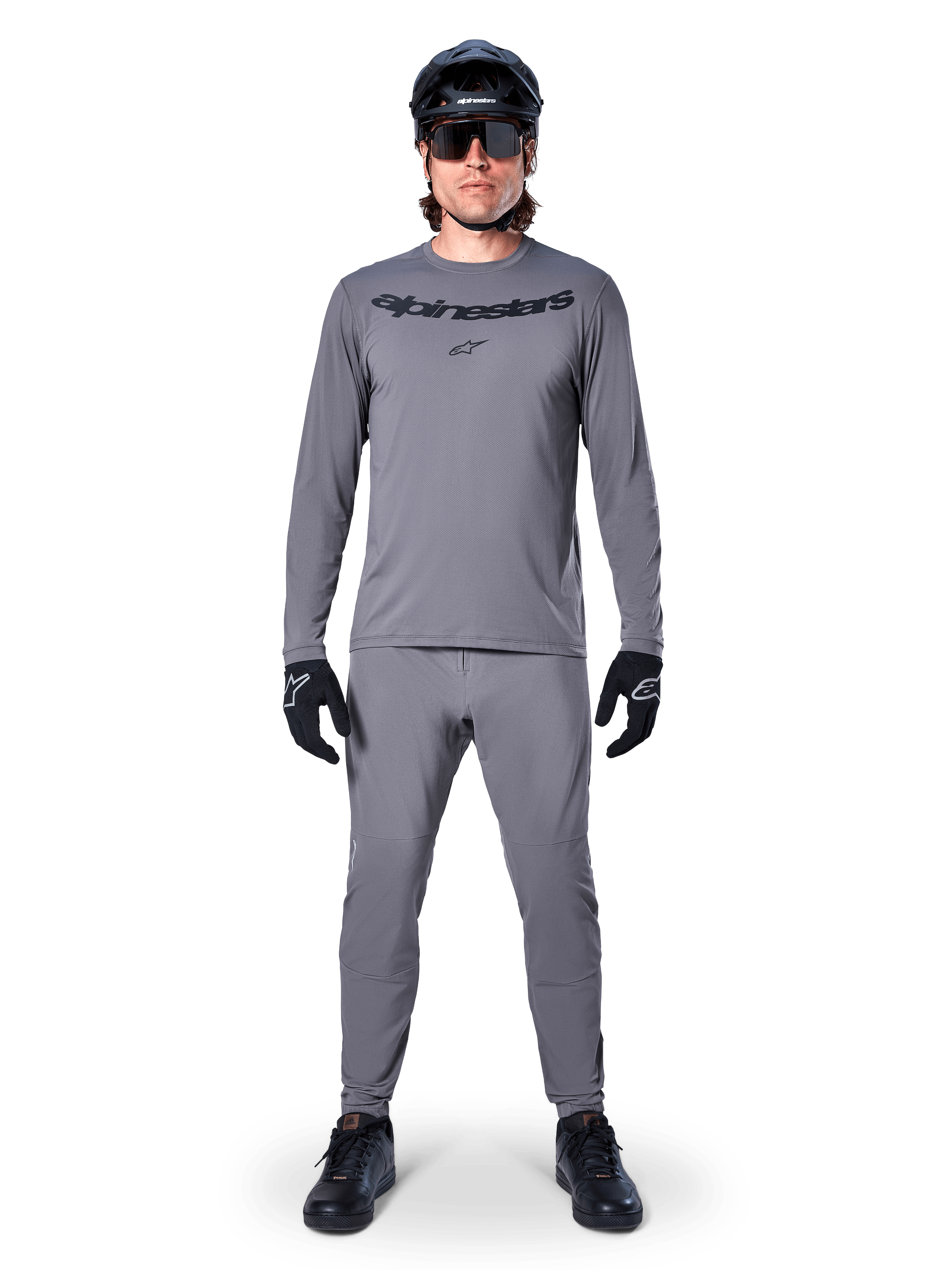 Kolarz w koszulce Alpinestars A-Dura Rocker z długim rękawem w kolorze ciemnoszarym, pasujące szare spodnie, czarny kask MTB, okulary przeciwsłoneczne i rękawiczki, profesjonalna odzież do jazdy na rowerze górskim na szlaki terenowe