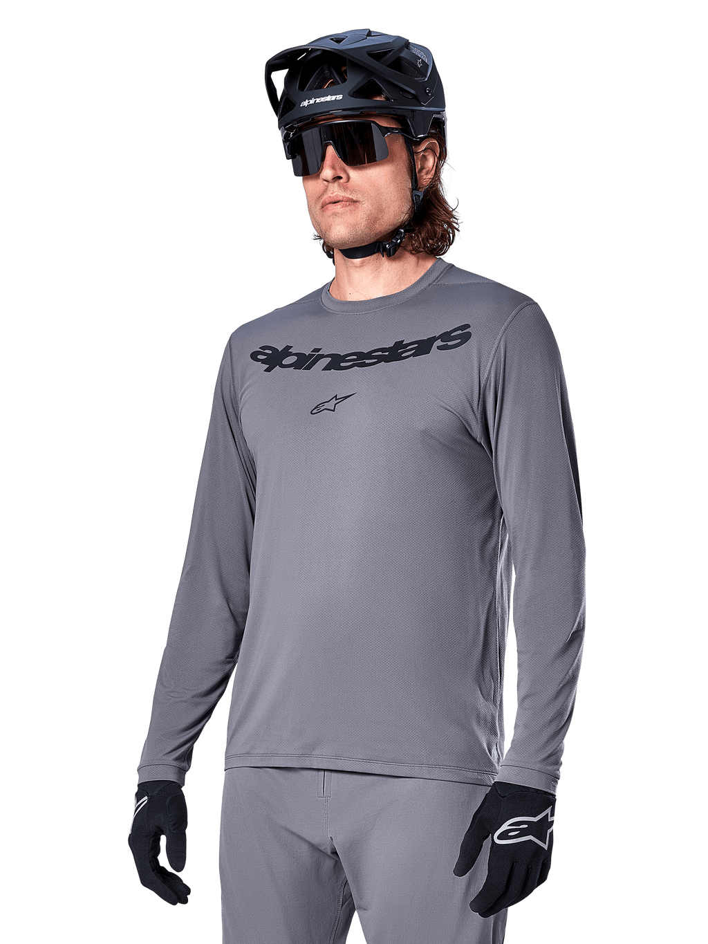 Alpinestars A-Dura Rocker Long Sleeve Jersey, szara koszulka MTB z długim rękawem, noszona z czarnym kaskiem i rękawicami, czarny napis na klatce piersiowej, sportowa odzież rowerowa do jazdy w terenie, jednolite tło