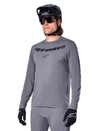 Alpinestars A-Dura Rocker Long Sleeve Jersey, szara koszulka MTB z długim rękawem, noszona z czarnym kaskiem i rękawicami, czarny napis na klatce piersiowej, sportowa odzież rowerowa do jazdy w terenie, jednolite tło