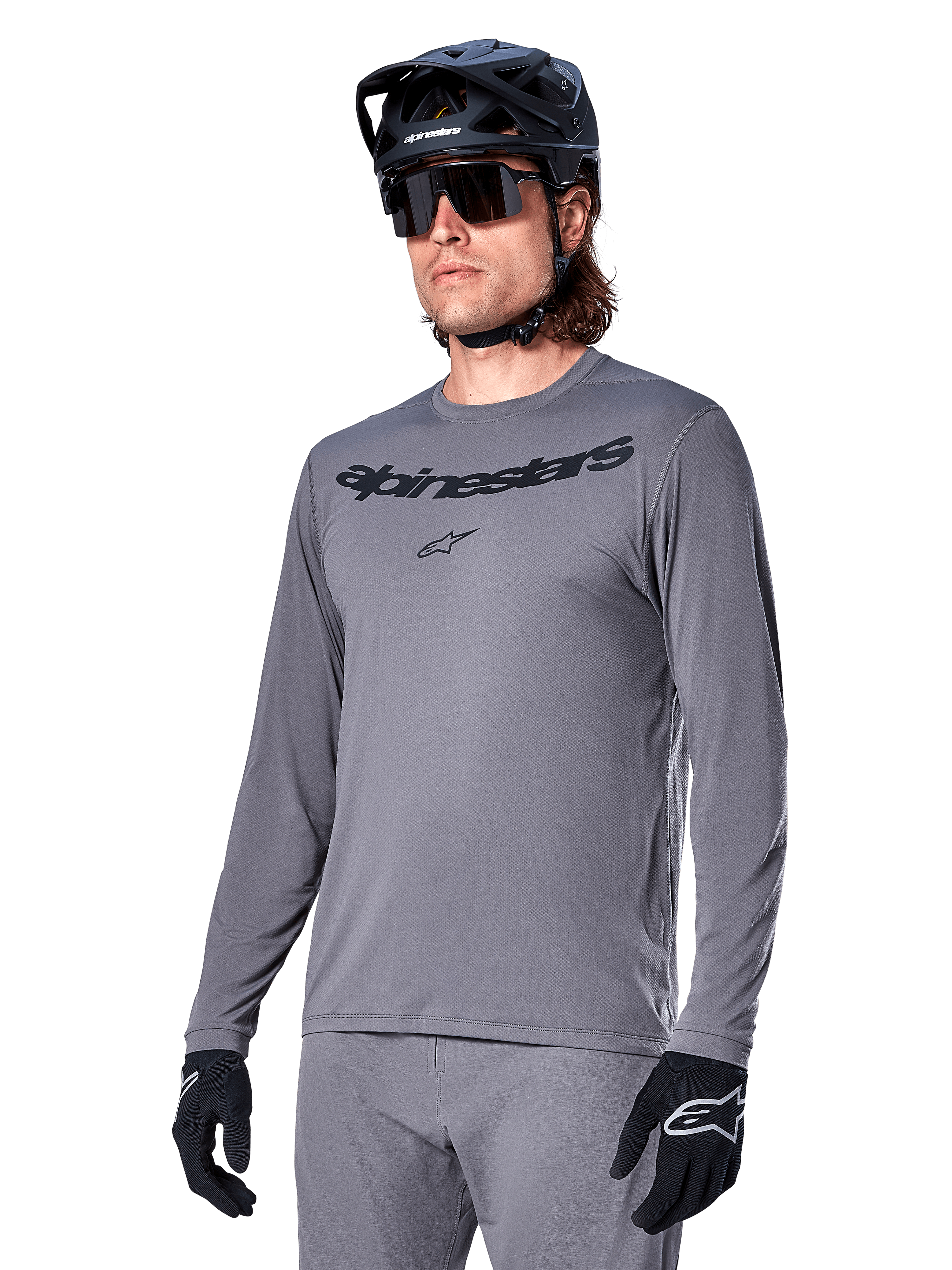 Alpinestars A-Dura Rocker Long Sleeve Jersey, szara koszulka MTB z długim rękawem, noszona z czarnym kaskiem i rękawicami, czarny napis na klatce piersiowej, sportowa odzież rowerowa do jazdy w terenie, jednolite tło