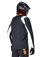 Rowerzysta MTB ubrany w koszulkę Alpinestars A-Supra Oscar z długim rękawem, czarna z białymi panelami na ramionach i rękawach, widok z tyłu, czarny kask i rękawice, spodnie z pomarańczowymi akcentami, wytrzymała odzież rowerowa off-road