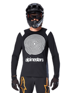 Alpinestars A-Supra Oscar Long Sleeve Jersey, czarna koszulka z długim rękawem, biała spiralna grafika i akcenty na ramionach, noszona przez rowerzystę w czarnym kasku full-face i goglach, czarno-pomarańczowe spodnie, wyczynowa odzież do jazdy MTB gravity