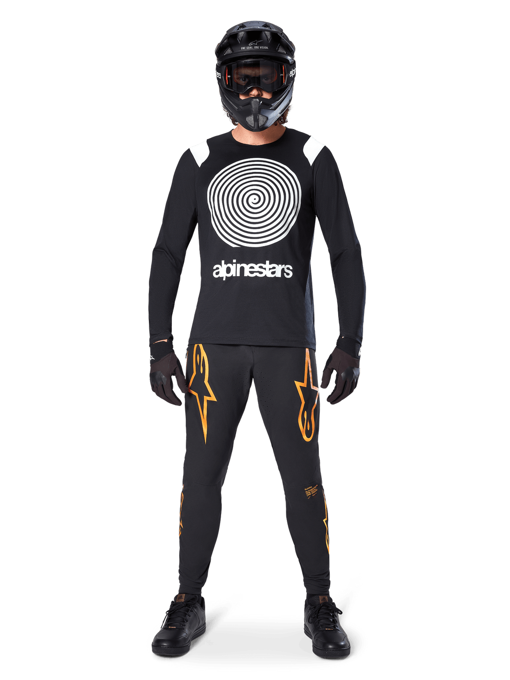 Męski model w kompletnym stroju MTB, koszulka Alpinestars A-Supra Oscar z długim rękawem, czarna z białą spiralną grafiką, czarne spodnie z pomarańczowym logo gwiazdy, czarny kask full-face i gogle, czarne rękawiczki i buty, dopasowany zestaw do jazdy grawitacyjnej