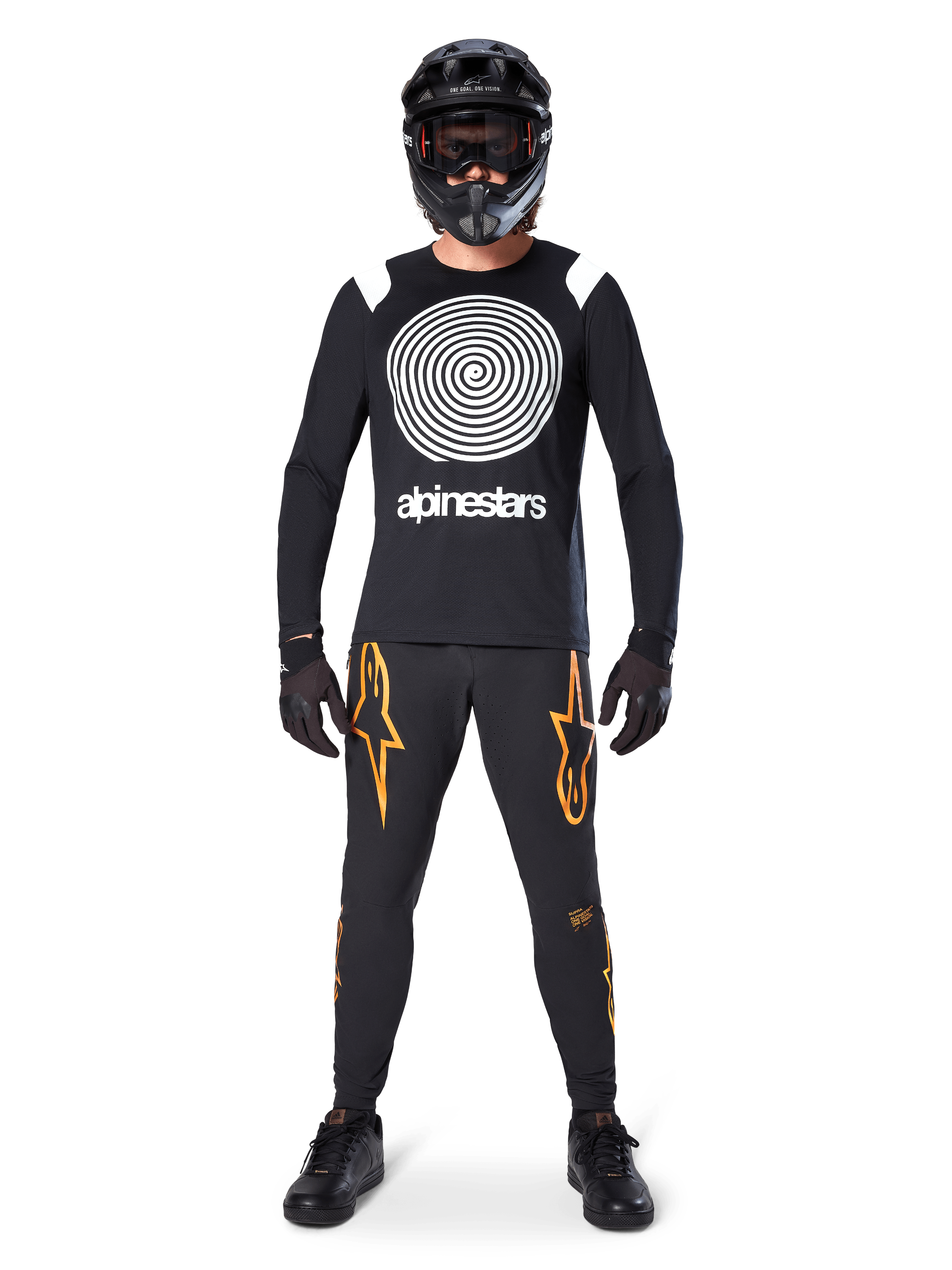 Męski model w kompletnym stroju MTB, koszulka Alpinestars A-Supra Oscar z długim rękawem, czarna z białą spiralną grafiką, czarne spodnie z pomarańczowym logo gwiazdy, czarny kask full-face i gogle, czarne rękawiczki i buty, dopasowany zestaw do jazdy grawitacyjnej