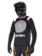 Alpinestars A-Supra Oscar Long Sleeve Jersey, koszulka rowerowa MTB, czarno-biała, z wyrazistym białym spiralnym wzorem i logo Alpinestars na klatce piersiowej, noszona z czarnym kaskiem full-face i goglami, profesjonalna odzież do kolarstwa górskiego
