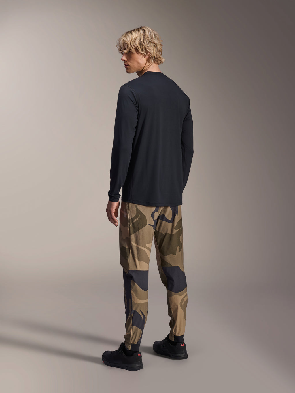 Alpinestars A-Dura Camo Pant, MTB Pants, Brown Camouflage, worn with a black long-sleeve jersey and black sneakers, rear view showing tapered legs and elasticated cuffs, performance apparel for off-road cycling