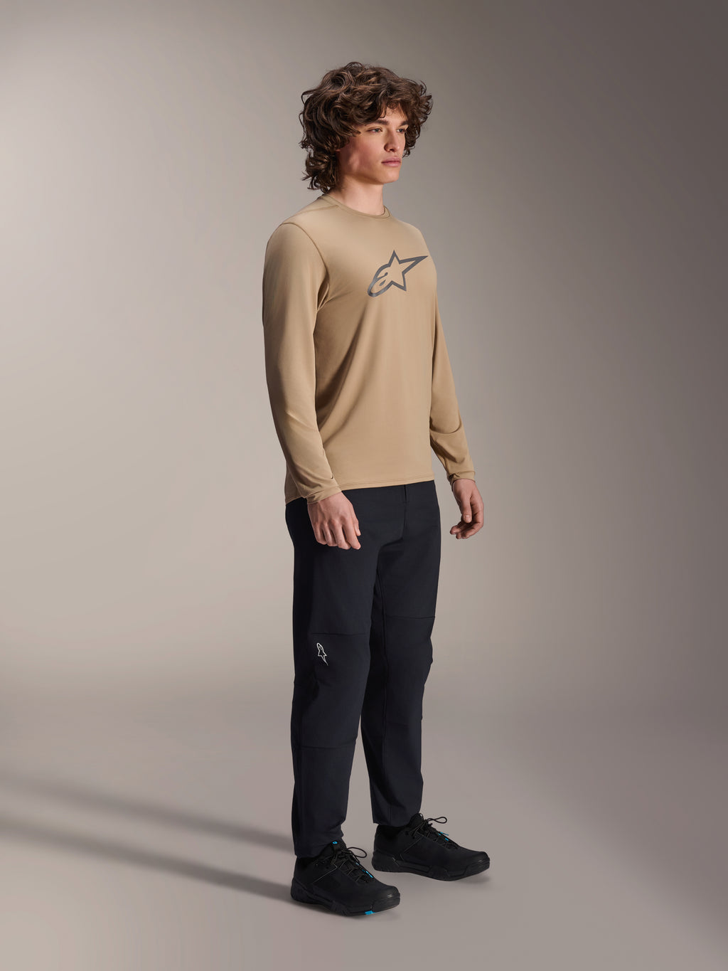 Alpinestars A-Dura Astar Long Sleeve Jersey, jasnobrązowa koszulka z długim rękawem, noszona przez modela z czarnymi spodniami i butami, szare logo Astars na piersi, casualowa odzież rowerowa MTB