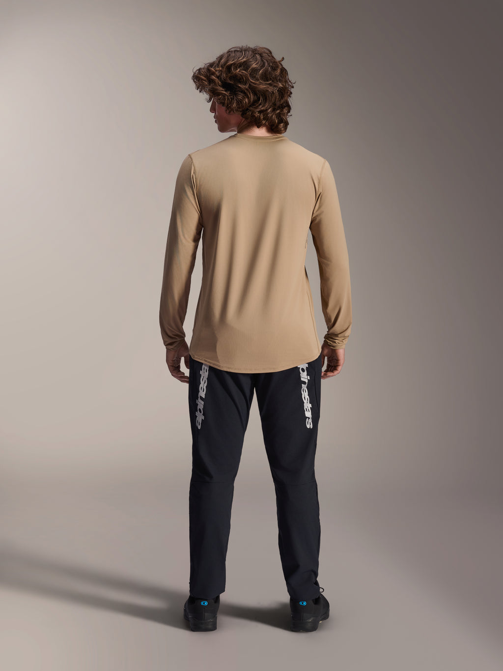 Alpinestars A-Dura Astar Long Sleeve Jersey, jasnobrązowa, widok z tyłu prezentujący przedłużony tył, noszona z czarnymi spodniami z brandingiem Alpinestars, odzież rowerowa MTB