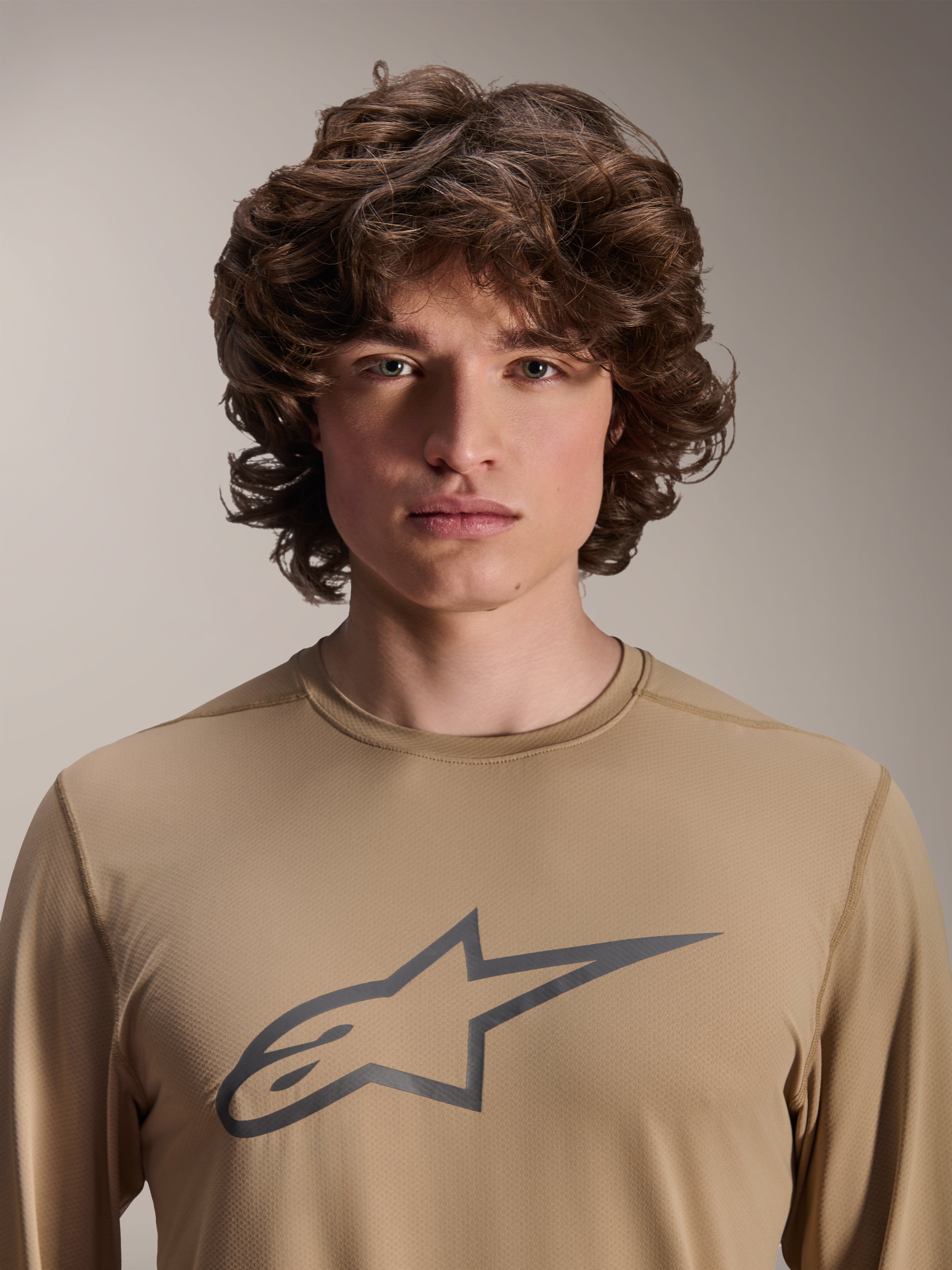 Alpinestars A-Dura Astar Long Sleeve Jersey, jasnobrązowa koszulka z długim rękawem, prezentowana przez modela, z dużym logo Astars na piersi, żakardowa tekstura materiału, casualowa odzież na rower górski