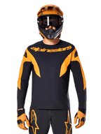 Rowerzysta MTB w koszulce Alpinestars A-Supra Race z długim rękawem, czarna z pomarańczową grafiką i logo na piersi, w zestawie z pomarańczowym kaskiem full-face, czarnymi goglami i pomarańczowymi rękawicami, profesjonalny strój do wyścigów górskich