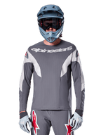 Rowerzysta MTB w koszulce Alpinestars A-Supra Race Jersey - Long Sleeve w kolorze ciemnoszarym, koszulka rowerowa z długim rękawem i białym logo Alpinestars, niebiesko-szary kask full-face, gogle i szare rękawice wyścigowe, kompletny strój wyścigowy do jazdy w terenie