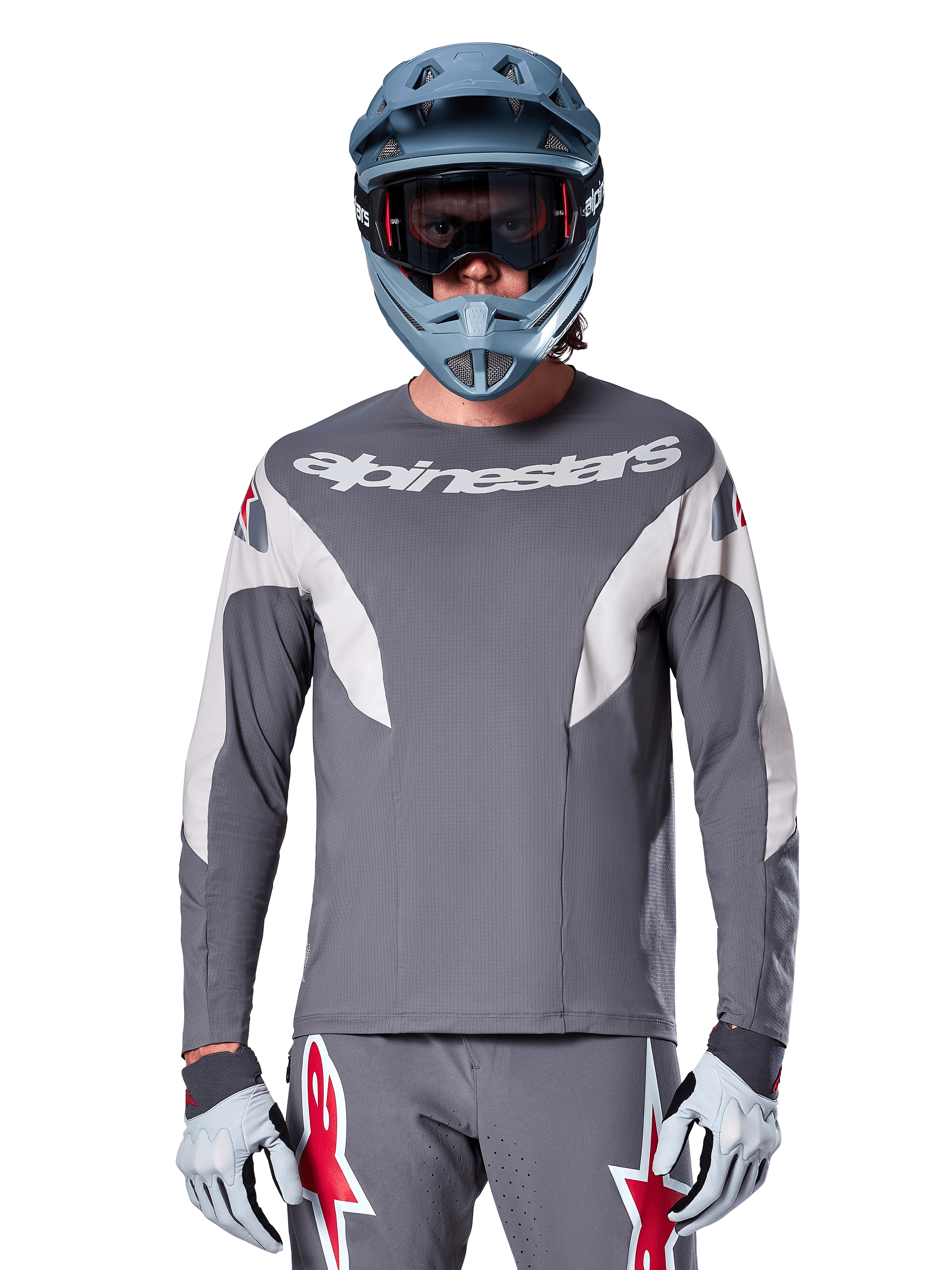 Rowerzysta MTB w koszulce Alpinestars A-Supra Race Jersey - Long Sleeve w kolorze ciemnoszarym, koszulka rowerowa z długim rękawem i białym logo Alpinestars, niebiesko-szary kask full-face, gogle i szare rękawice wyścigowe, kompletny strój wyścigowy do jazdy w terenie