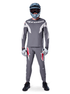 Rowerzysta w koszulce Alpinestars A-Supra Race Jersey z długim rękawem w kolorze ciemnoszarym, zestawionej z dopasowanymi szarymi spodniami z wyrazistymi czerwonymi logotypami gwiazdy, szarym kaskiem z goglami i jasnoszarymi rękawicami; kompletny zestaw wyścigowy MTB do wyczynowej jazdy terenowej