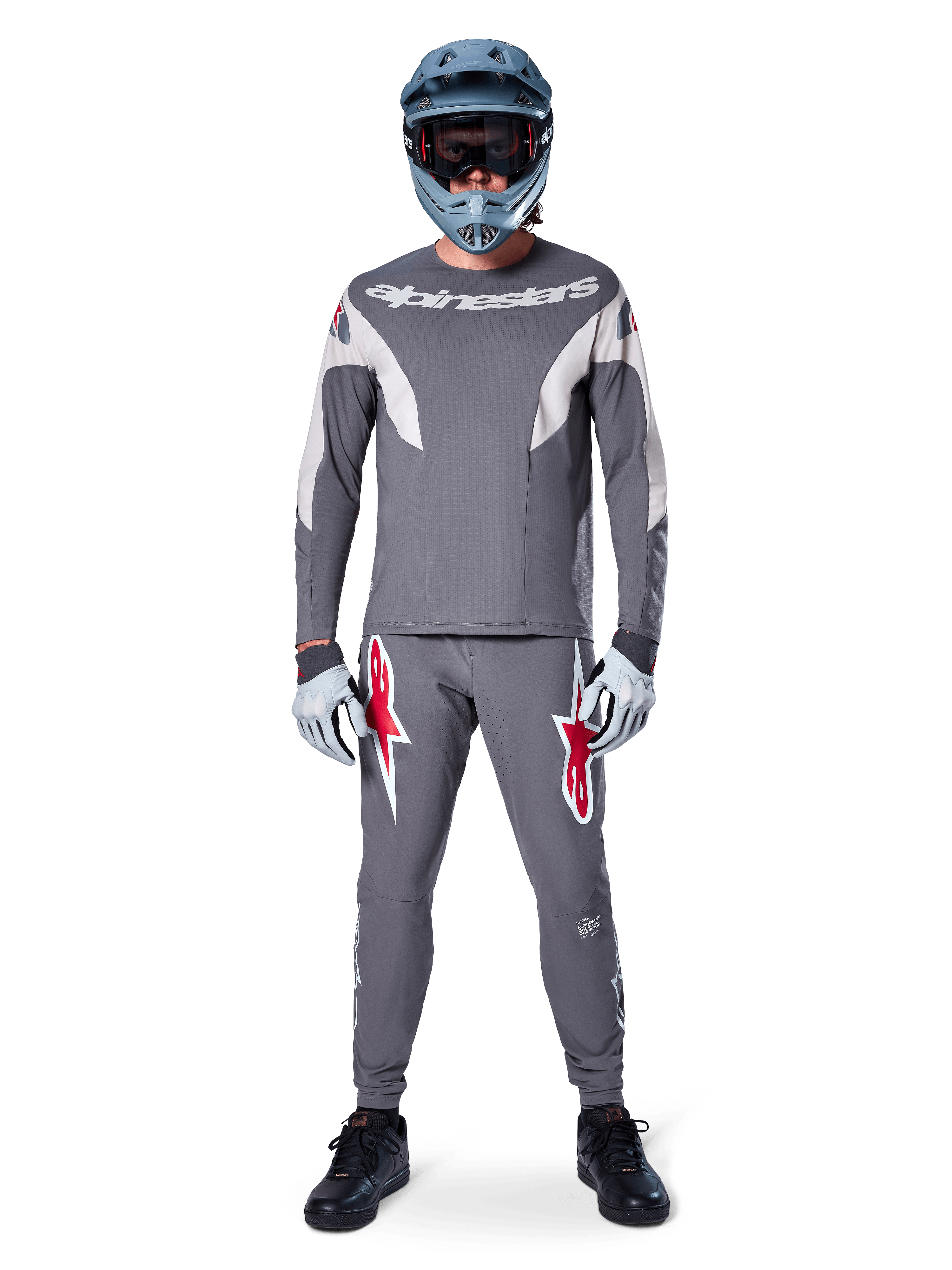 Rowerzysta w koszulce Alpinestars A-Supra Race Jersey z długim rękawem w kolorze ciemnoszarym, zestawionej z dopasowanymi szarymi spodniami z wyrazistymi czerwonymi logotypami gwiazdy, szarym kaskiem z goglami i jasnoszarymi rękawicami; kompletny zestaw wyścigowy MTB do wyczynowej jazdy terenowej