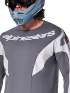 Alpinestars A-Supra Race Jersey - Long Sleeve, koszulka MTB, ciemnoszara z białą grafiką wyścigową i białym logo Alpinestars, noszona z jasnoniebieskim kaskiem full-face i goglami, mikroperforowany materiał dla wydajności off-road