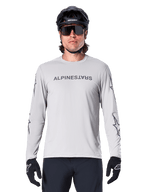 Alpinestars A-Dura Switch Long Sleeve Jersey, jasnoszara, posiada ciemnoszary napis na klatce piersiowej i logotypy gwiazdy na rękawach, noszona przez rowerzystę w czarnym kasku MTB i rękawiczkach, wydajna odzież rowerowa do jazdy po szlakach
